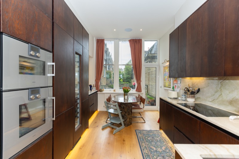 Galley kitchen with dark walnut cabinetry, integrated appliances and dining nook beneath a sash bay window