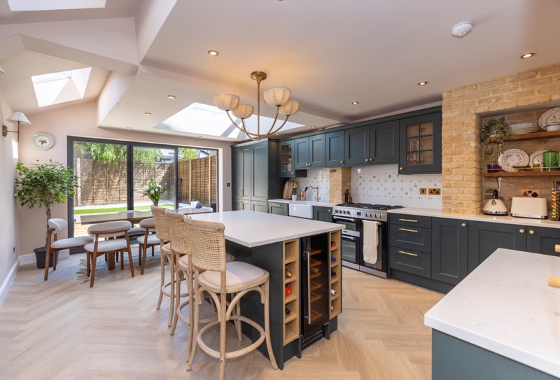 Dark green Shaker kitchen with marble island, exposed brick alcove and open shelving beneath rooflight