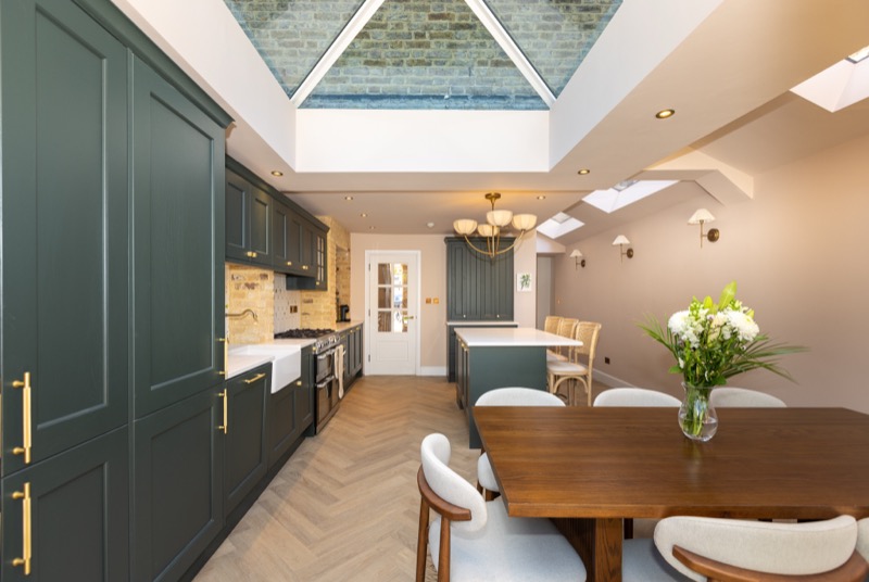 Galley view of a green Shaker kitchen with pyramid skylight and walnut dining table beyond